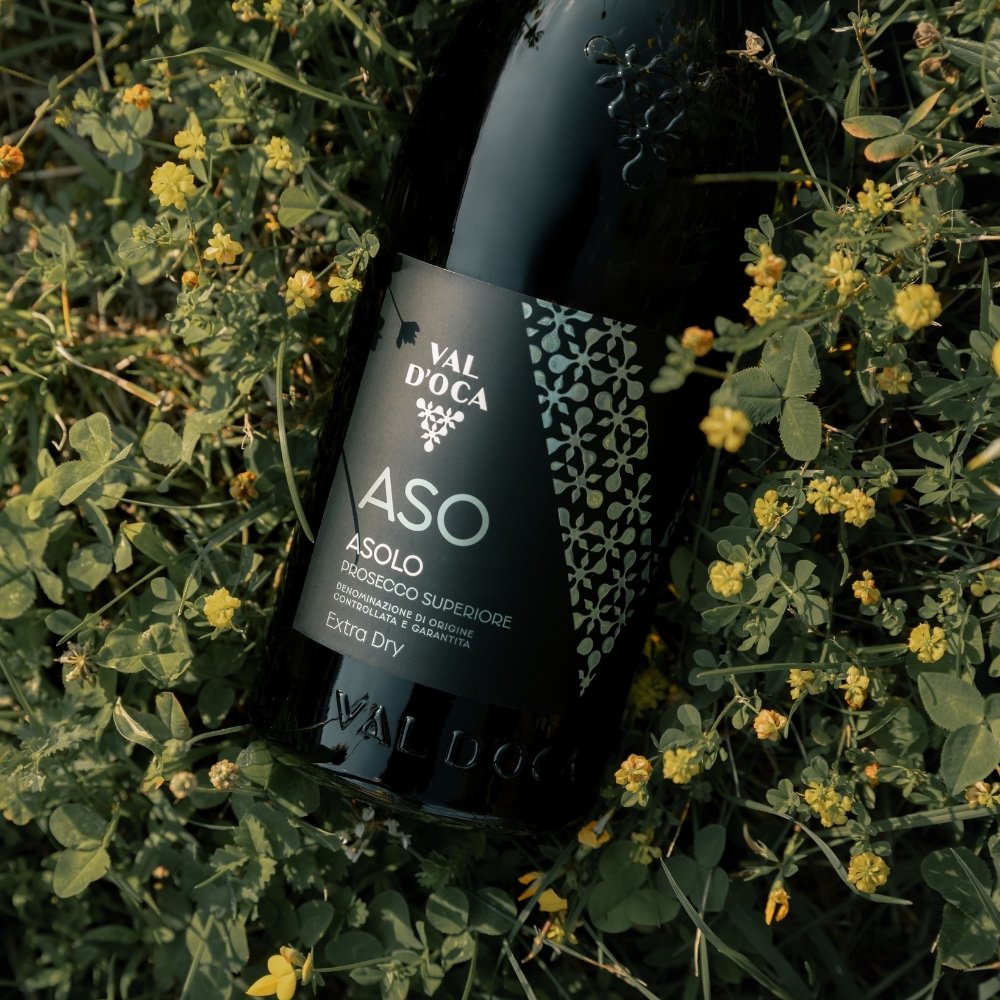 Bottiglia scura di Val d'Oca ASO Asolo Prosecco Superiore Extra Dry adagiata tra trifoglio e fiori gialli